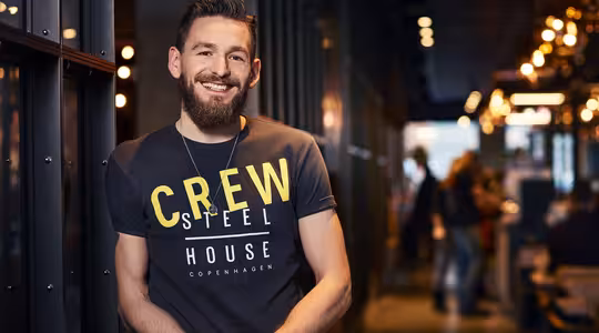 En man med skägg ler och lutar sig mot en mörk vägg i en varmt upplyst hall. Han har på sig en t-shirt där det står "Crew Steel House Copenhagen". Bakgrunden har suddiga ljus och människor.