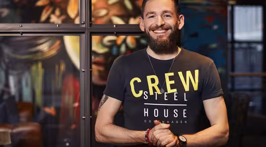 Leende man med skägg står i ett färgglatt rum. Han har på sig en svart T-shirt med orden "CREW STEEL HOUSE COPENHAGEN" tryckta med gula och vita bokstäver. Han har en klocka och rött armband på handleden.