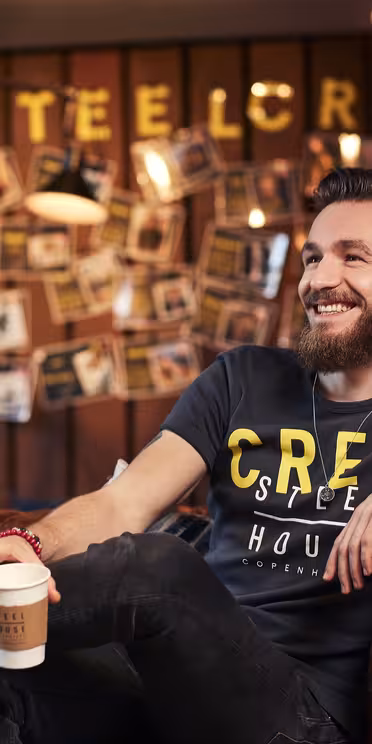 En skäggig man sitter på en brun skinnsoffa och ler och håller i en kaffekopp. Han bär en svart t-shirt och sneakers. Bakgrunden visar bilder och ordet "#SteelCrew" med upplysta bokstäver. Miljön är mysig och varmt upplyst.