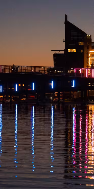 En bro upplyst med färgglada LED-lampor reflekteras på vattenytan i skymningen. Moderna byggnader och kranar i bakgrunden siluetteras mot solnedgångshimlen, vilket bidrar till det urbana landskapet.