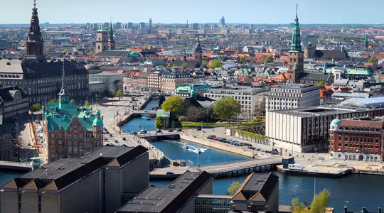 Flygfoto över Köpenhamn, som visar upp historisk och modern arkitektur med kanaler som skär genom staden. Framstående byggnader och kyrktorn reser sig ovanför, omgivna av träd och urban infrastruktur under en klarblå himmel.