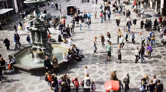 Ett livligt europeiskt torg med människor som går och samlas. Det finns en fontän till vänster och historiska byggnader med olika fasader kantar gatorna. Kullerstensmönster täcker marken och atmosfären är livlig och levande.