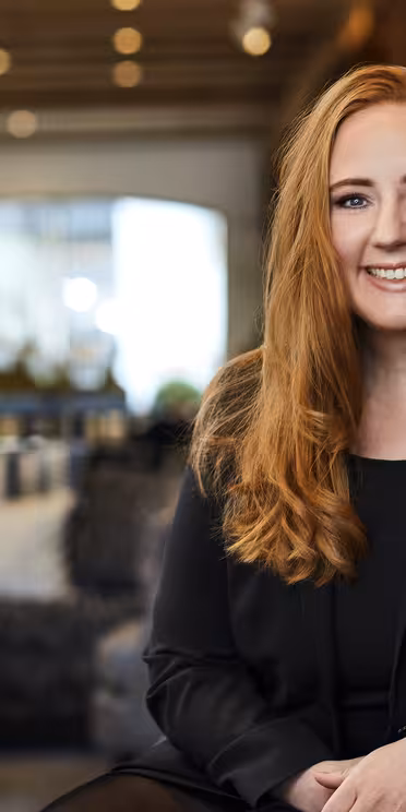 Leende kvinna med långt rött hår klädd i en svart kavaj och en namnlapp, sitter i en mysig, modern lounge med mjuk belysning och suddig bakgrund.