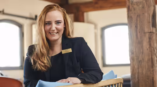 En kvinna med långt rött hår ler, iklädd en svart kavaj och en namnskylt. Hon sitter inomhus framför rustika träbjälkar och mjuk belysning, med två fönster i bakgrunden.