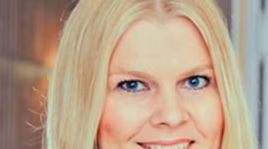 En person med långt blont hår, ler, befinner sig i ett väl upplyst rum. De bär en blå topp, och den suddiga bakgrunden visar en trävägg och ett mjukt ljus.