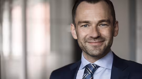 En man i mörkblå kostym och randig slips ler mot kameran och står i ett mjukt upplyst rum med suddig bakgrund.