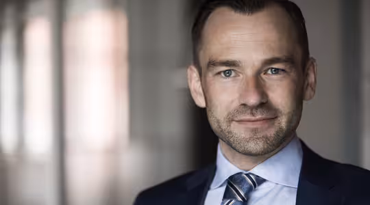 En man i kostym med kort hår och ljust skägg ler lätt mot en suddig kontorsbakgrund. Han är klädd i blå slips och kostymjacka, med vit skjorta. Belysningen är mjuk och framhäver hans drag.