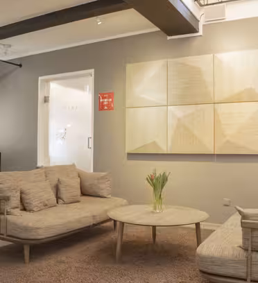 En modern, minimalistisk lounge har två beiga soffor, ett runt soffbord med en vas med tulpaner och ett geometriskt träkonstverk på en grå vägg. Utrymmet har en grå gardin och en dörr som leder till ett annat rum.