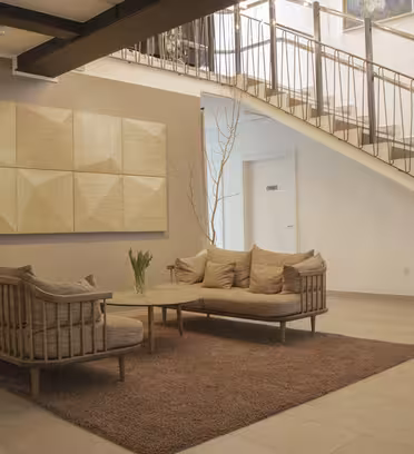 En modern lobby med ett soffbord i trä och två vadderade soffor placerade på en brun matta. Ett geometriskt konstverk hänger på väggen. En trappa med metallräcken syns i bakgrunden. En liten vas med blommor står på bordet.