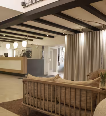 Modern hotellobby med beiga soffor och en stor vas. En receptionsdisk i trä finns i bakgrunden, bredvid en spiraltrappa. I taket hänger taklampor och en skir gardin delar upp utrymmet.