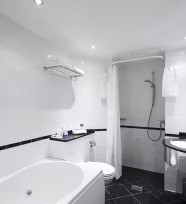 Modernt badrum med vita kaklade väggar och svart klinkergolv. Har ett stort badkar, en hörndusch med gardin, en toalett och ett handfat med en svart bänkskiva. Handdukar hänger från ställ och utrymmet är väl upplyst med taklampor.