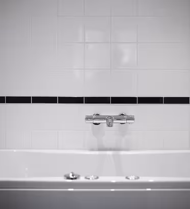 Ett vitt badkar med en silverblandare mot en vägg av vitt kakel, med en enda rad mörka kakel som accent. Miljön är minimalistisk och modern, med mjuk belysning.