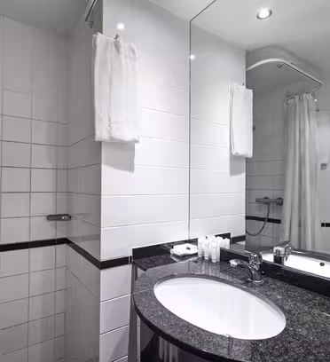 Modernt badrum med vita kaklade väggar och svarta detaljer. Har en bänkskiva i svart granit med oval handfat och kromblandare. En stor spegel är ovanför diskbänken. Duschdelen har en glaspanel och ett silverduschhuvud med vit gardin.