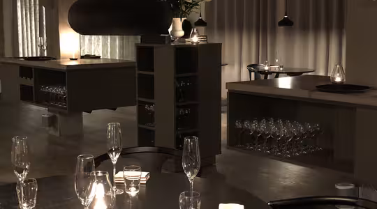 En svagt upplyst, modern restaurang med runda bord för middagar. Varje bord har tomma vinglas, små ljus och vikta servetter. Taklampor ger ett varmt sken. Hyllor med glasögon och gardiner syns i bakgrunden.