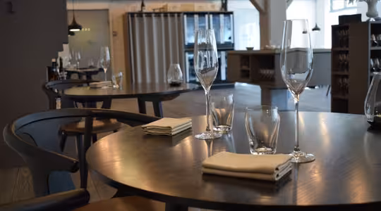 En modern, elegant matplats med runda träbord med vikta servetter, tomma vinglas och vattenglas. Mjuk belysning skapar en varm atmosfär. I bakgrunden förstärker hyllor och armaturer den moderna inredningen.