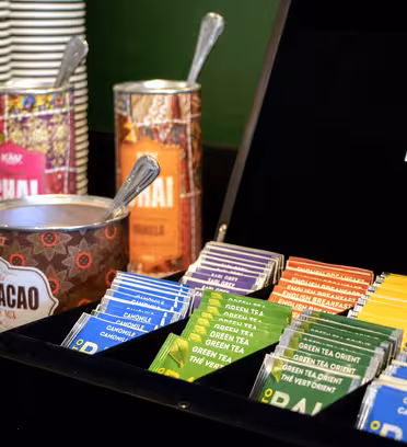 Ett sortiment av tepåsar organiserade i en svart låda, med olika smaker som kamomill, grönt te och chai. Bakom lådan står staplade pappersmuggar, en skål med sockerpaket och burkar märkta "CACAO" och "CHAI.