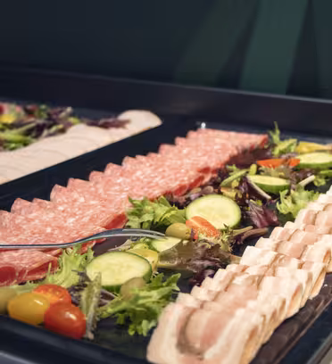 En buffétallrik med ett sortiment av skivade delikatesser, inklusive salami och kalkon, garnerad med blandade grönsaker, körsbärstomater och gurkskivor.