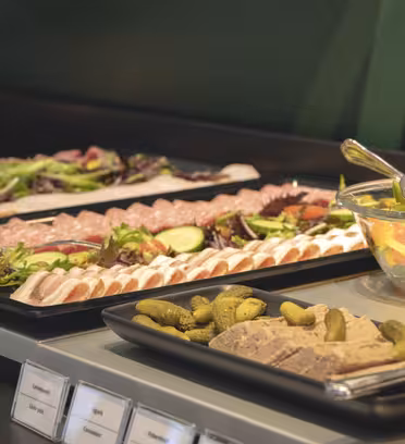 Ett buffébord med brickor med diverse delikatesskött, skivade gurkor, körsbärstomater och bladgrönsaker. En skål med färgglad fruktsallad och ett fat med små pickles visas också. Etiketter är synliga framför brickorna.