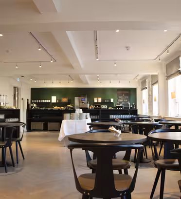 En modern caféinteriör med runda mörka bord och trästolar. I bakgrunden finns en serveringsyta med en kaffemaskin, staplade tallrikar och matvaror. Stora fönster släpper in naturligt ljus och lyser upp det ljusa, rymliga rummet.