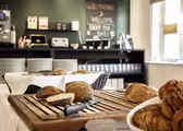 Bröd och bakverk visas på en skärbräda i trä i ett mysigt kök med mörkgrön vägg. En svart tavla i bakgrunden läser, "god morgon & välkomnande. Njut av din dag!" Tallrikar och kaffebryggare syns på hyllorna.