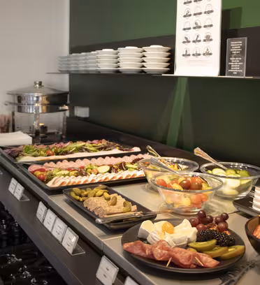 Ett buffébord med olika matvaror, inklusive skivat kött, ostar, färsk frukt, salladsskålar och små tallrikar, mot en grön och svart vägg med extra tallrikar staplade på en hylla ovanför.