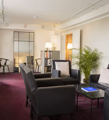 En modern lounge med svarta läderstolar och en soffa på en rödbrun matta. Rummet har inramad abstrakt konst, en stor krukväxt, en bokhylla och en hög spegel. Mjuk belysning och randiga gardiner skapar en inbjudande atmosfär.