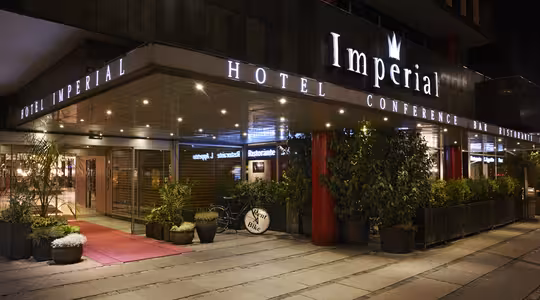Gatuvy över entrén till Hotel Imperial. Byggnaden har upplyst skyltning och ett tak med krukväxter längs gångvägen. En röd matta leder till entrédörrarna som ramas in av stora fönster som visar innerbelysning.