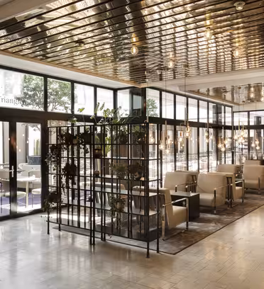 Modern hotelllounge med fönster från golv till tak, avdelade sittgrupper med beiga stolar och bord samt dekorativ grönska. Taket har reflekterande metallpaneler och golvet är lättkaklat.