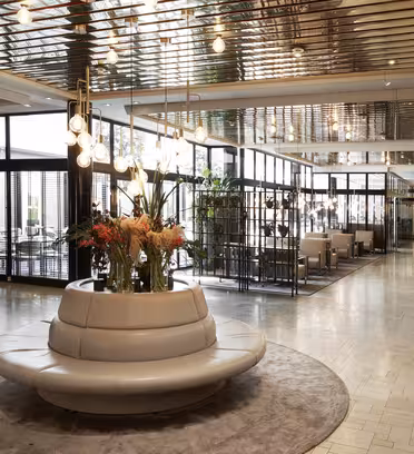 Modern hotellobby med en rund grå soffa centrerad på en rund matta. Ett blomsterarrangemang och taklampa ovanför soffan. Glasväggar separerar mötesrum till höger med en stor krukväxt till vänster.