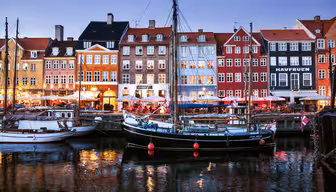En naturskön utsikt över Nyhavn i Köpenhamn i skymningen, med färgglada historiska byggnader och förtöjda båtar som reflekteras i det lugna kanalvattnet. Området är upplyst med mjukt ljus från kaféerna och restaurangerna vid vattnet.