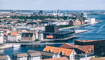 Flygfoto över en kuststad med en blå vattenväg och bryggor. Stadsbilden har en blandning av moderna och historiska byggnader med röda tegeltak. En stor, modern byggnad är framstående längs vattnet. Himlen är klar med några moln.
