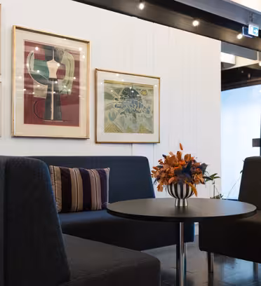 Modern kontorslounge med två mörkgrå stolar, ett runt svart bord med en vas med orange blommor. Väggarna har inramad abstrakt konst. Utrymmet är väl upplyst, med en blandning av naturligt och artificiellt ljus.
