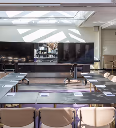 Ett ljust konferensrum med två stora skärmar framtill, arrangerade i U-form med bord och beiga stolar. Naturligt ljus strömmar in genom ett takfönster och växter syns i bakgrunden. Anteckningsblock läggs på borden.