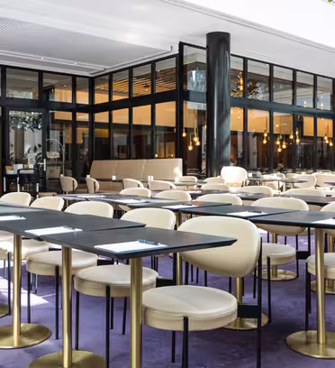 Modern restauranginteriör med bord och beige stolar ordnade prydligt i rader. Mjuka lila heltäckningsmattor och stora fönster släpper in naturligt ljus i rummet. Anteckningar och pennor placeras på varje bord, vilket skapar en konferens- eller mötesuppställning.