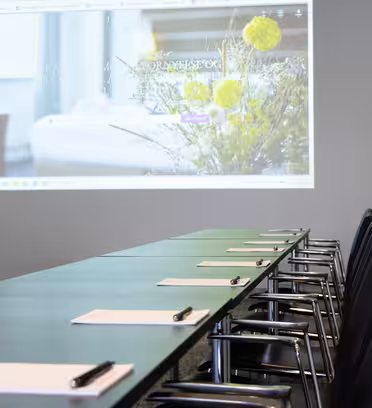 Ett långt konferensbord med svarta stolar, var och en uppsättning med ett anteckningsblock och penna, vetter mot en skärm som projicerar en suddig webbplats med blommiga bilder. Rummet är heltäckningsmatta och väggarna är enkla, vilket skapar en modern, professionell mötesplats.