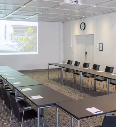 Ett modernt, tomt konferensrum med U-formade svarta bord, svarta stolar och en presentationsskärm som visar en naturbild. Rummet har en grå matta, vita väggar och en klocka som visar 12:05 ovanför en stängd dörr.