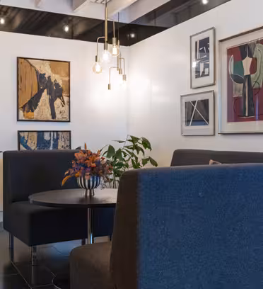 Ett modernt, elegant inrett rum med två mörkgrå soffor runt ett runt svart bord med en blommig mittpunkt. Konstverk visas på vita väggar och hängande glödlampor lyser upp utrymmet. En växt placeras nära en vägg.