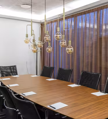Ett modernt konferensrum med ett långt träbord omgivet av tio svarta läderstolar. Anteckningsblock och pennor placeras framför varje stol. Över huvudet hänger dekorativa taklampor och i bakgrunden finns ett stort fönster med bruna gardiner.