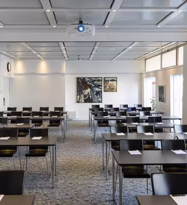 Ett tomt konferensrum med rader av bord och stolar i klassrumsstil. En projektor är monterad i taket och anteckningsblock placeras på varje bord. Två målningar finns på bakväggen. Stora fönster kantar rummets högra sida.