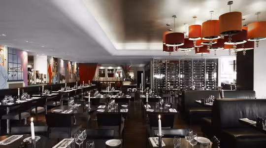 Exklusiv restauranginteriör med svag belysning, snyggt dukade bord med ljus och glas. Modern inredning inkluderar röda ljuskronor, konstverk på väggarna och ett stort vinställ. Atmosfären är elegant och inbjudande.