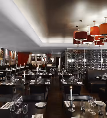 Modern restauranginteriör med bord och stolar i mörkt trä, svarta läderstolar och röda trumljuskronor. Borden är dukade med glas och ljus. En vinutställning och färgglad väggkonst bidrar till den eleganta atmosfären.