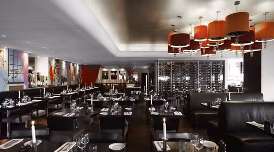 Modern restauranginteriör med bord och stolar i mörkt trä, svarta läderstolar och röda trumljuskronor. Borden är dukade med glas och ljus. En vinutställning och färgglad väggkonst bidrar till den eleganta atmosfären.