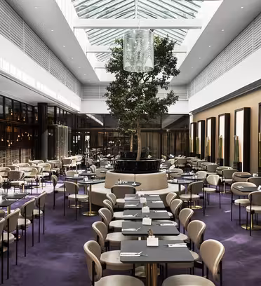 En rymlig modern restaurang med högt i tak och stora takfönster. Den har ett centralt träd, beiga stolar, svarta bord och dekorativa väggar med upplysta paneler. Mattan är lila och inredningen är minimalistisk och elegant.