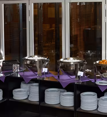 En elegant bufféuppsättning i ett svagt upplyst rum. Flera skavskålar rymmer olika livsmedel. Vita tallrikar är snyggt staplade nedanför. Lila servetter framhäver bordet. Stora fönster med träpersienner i bakgrunden.