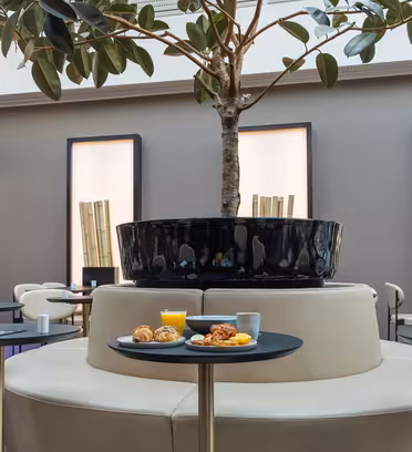 En modern matplats med runda bord med stolar, centrerad runt ett stort krukträd. Tallrikar med bakverk och ett glas apelsinjuice står på ett bord. Inredningen inkluderar höga bambuliknande installationer mot en minimalistisk bakgrund.