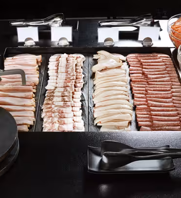 Ett urval av delikötter visas på en svart bänkskiva, inklusive remsor av bacon, kyckling och korv. Skålar med skivade gurkor, tomater och paprika ligger vid sidan av. Svart tång och småfat står framme.