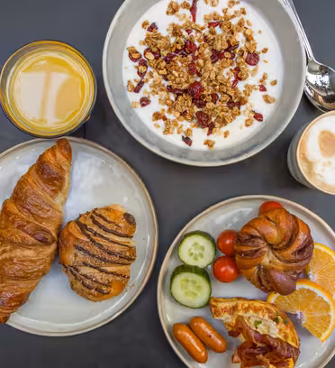 En frukost som serveras på ett bord inkluderar en skål med yoghurt med granola, ett glas apelsinjuice, en kopp cappuccino, croissanter, ett bakverk, skivade apelsiner, gurka, körsbärstomater, minikorvar och bacon.