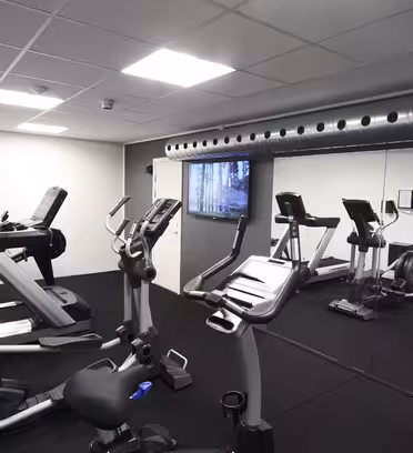 Modernt gym med träningsutrustning, inklusive löpband, stationära cyklar och elliptiska maskiner. Rummet har spegelvägg, svart golv och tak med infällda lampor och synliga kanalsystem.