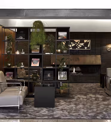 Modern lounge med grå fåtöljer och en geometrisk hyllenhet som visar växter och konst. En öppen spis finns i bakgrunden och träpaneler ger värme. Det heltäckningsmatta golvet och den minimalistiska inredningen skapar en mysig atmosfär.