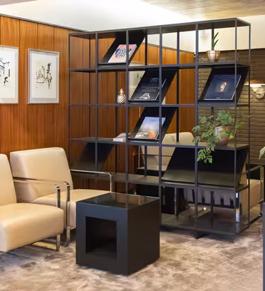Modern loungedel med två beiga fåtöljer och ett svart fyrkantigt bord. En geometrisk svart hylla visar böcker och dekorativa föremål. Väggen är prydd med abstrakt konst. En stor växt sitter i en vas på höger sida. Golvet är heltäckningsmatta.